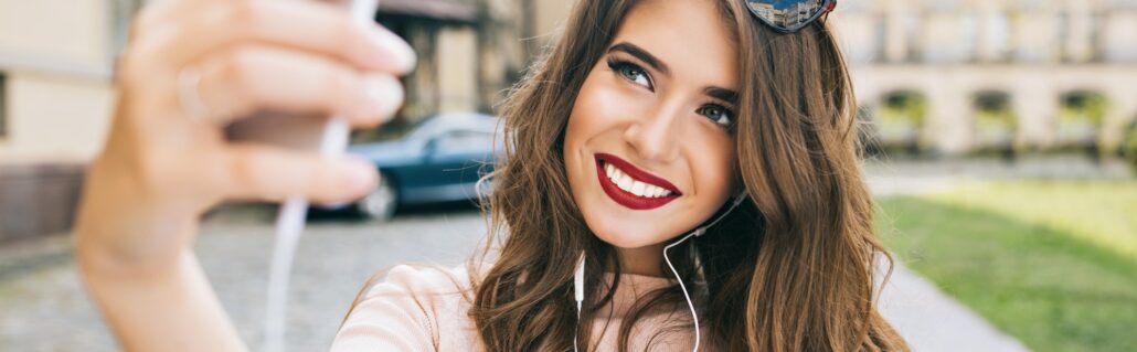 Portrait of cute girl with long hair and snow-white smile making selfie on street in city. She wears